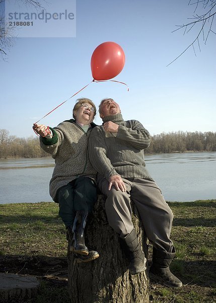 Seniorenpaar am Fluss sitzend mit rotem Ballon  lächelnd