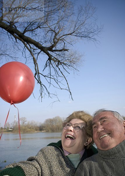 Seniorenpaar am Fluss hält roten Ballon  Köpfe zusammen  lächelnd