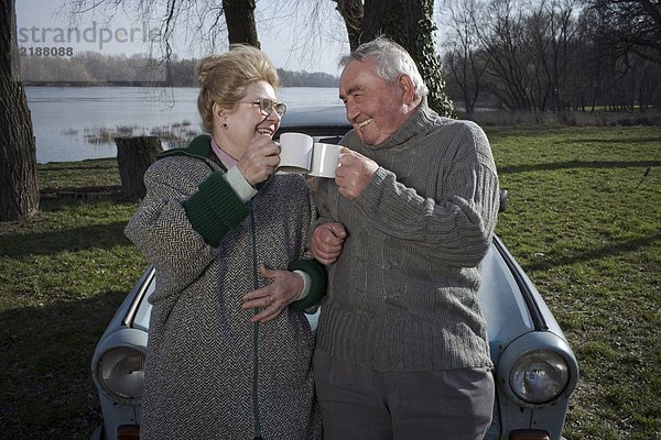 Seniorenpaar steht neben dem Auto und toastet sich gegenseitig mit Tassen  lächelt.