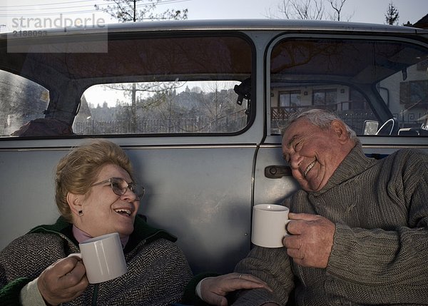 Seniorenpaar sitzt mit dem Auto auf dem Land  hält Tassen  lächelt