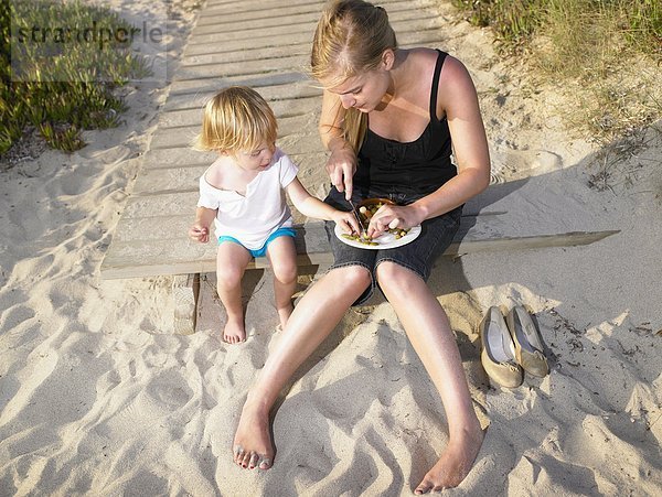 Frau sitzt mit einem kleinen Jungen am Strand und isst.