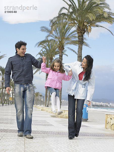 Mutter und Vater gehen auf einem palmengesäumten Bürgersteig am Meer entlang und schwingen die kleine Tochter (6-8). Alicante  Spanien.