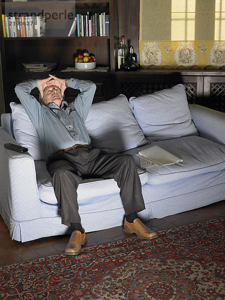 Älterer Mann  der die Stirn auf einem Sofa hält  das vom Fernsehen beleuchtet wird.