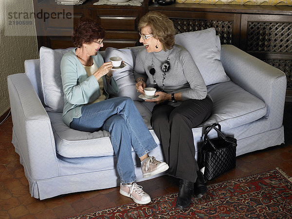 Zwei ältere Frauen  die auf dem Sofa sitzen und Tassen halten und lachen.