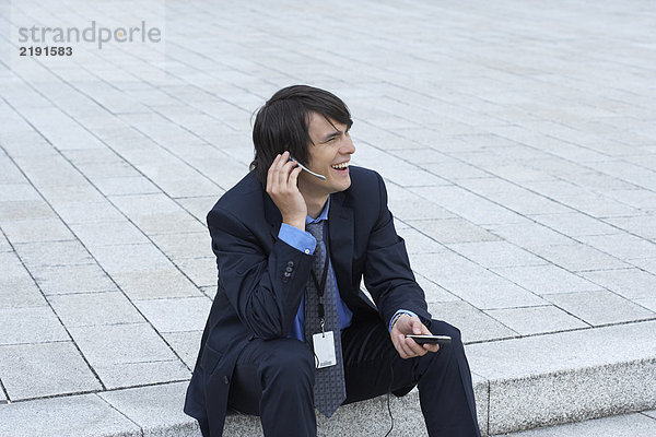 Geschäftsmann auf einer Treppe sitzend  mit Kopfhörer und mobilem Gespräch  lächelnd in die Ferne blickend.