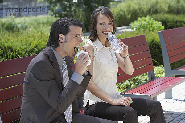Geschäftsmann und Frau  die draußen auf einer Bank sitzen und Wasser trinken wollen.