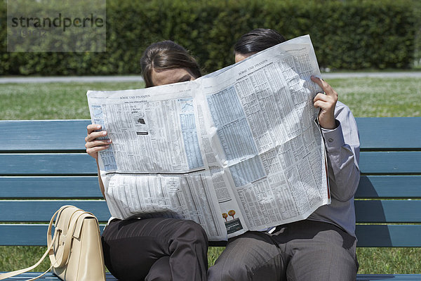 Geschäftsmann und Frau auf der Bank im Park  die zusammen eine Zeitung lesen  die sich dahinter versteckt.