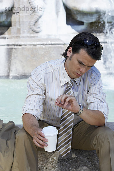 Geschäftsmann sitzt am Brunnenrand mit Kaffee  um auf seine Uhr zu schauen.