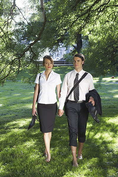 Junges Paar hält sich an den Händen und geht barfuß auf der Wiese spazieren.