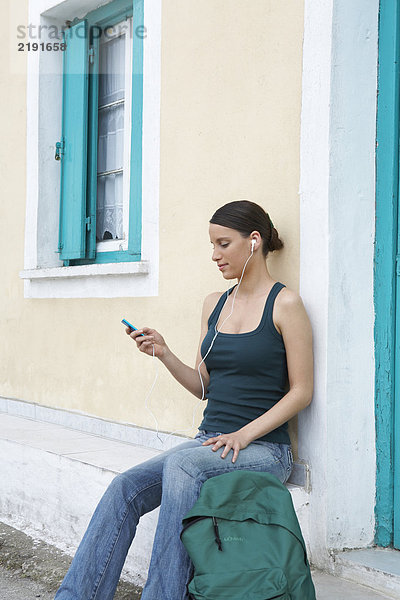 Junge Frau sitzt vor einem griechischen Haus mit Rucksack und Kopfhörern und schaut auf das MP3-Display.