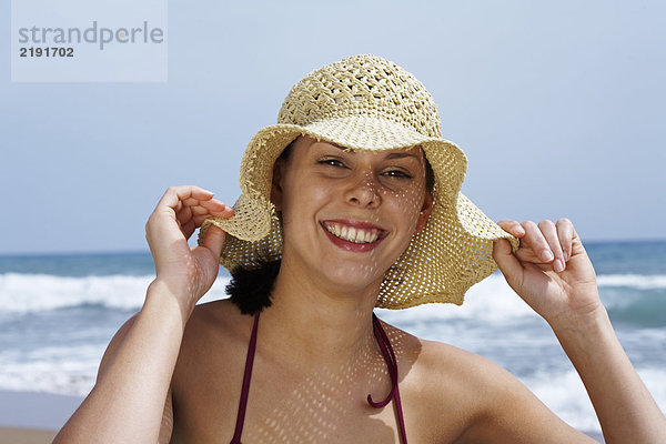 Junge Frau im Bikini am Strand mit Strohhut und Blick ins Meer im Hintergrund.