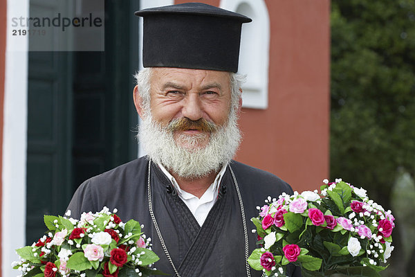 Alter Priester vor der Kirche mit zwei Blumensträußen  die lächelnd in die Kamera schauen.