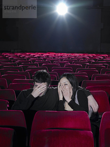 Junges Paar sitzt im Kino und hält die Hände über die Gesichter.