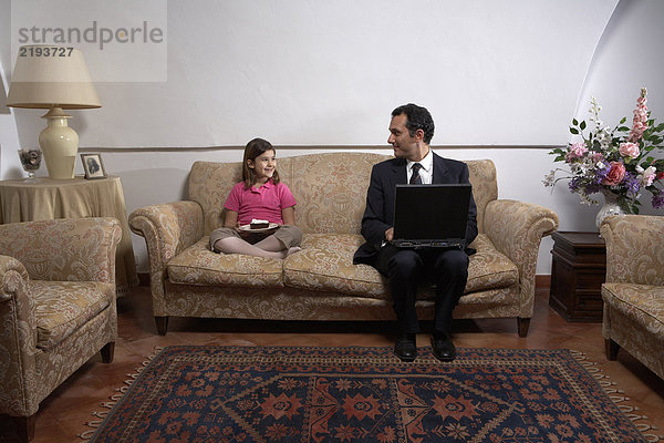 Mann mit Laptop mit Blick auf Tochter (6-8) auf Sofa mit Kuchen