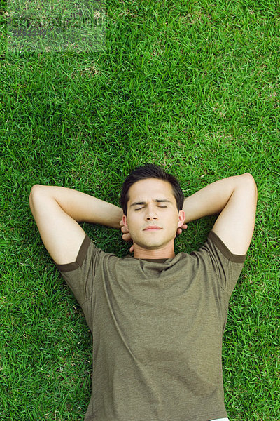 Junger Mann auf Gras liegend  Hände hinter dem Kopf  Augen geschlossen  Hochwinkelansicht