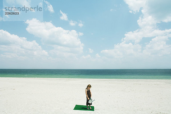 Mädchen steht auf Kunstrasenplatz am Strand  hält Gießkanne  Blick in den hohen Winkel