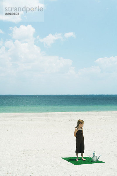 Mädchen steht auf Kunstrasenplatz am Strand  neben der Gießkanne  Blick in den hohen Winkel