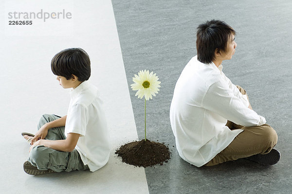 Junge und Mann sitzen Rücken an Rücken  eine einzige Blume zwischen ihnen.
