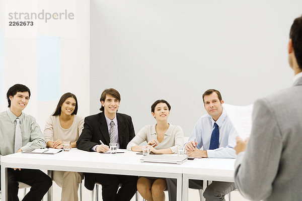 Gruppe von Profis am Tisch sitzend  lächelnd  mit Blick auf den Mann im Vordergrund.