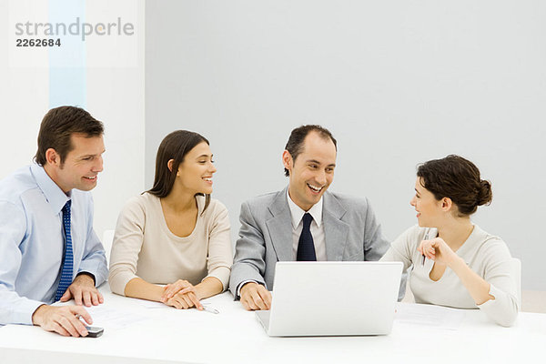 Gruppe von männlichen und weiblichen Geschäftspartnern  die zusammen am Tisch mit einem Laptop sitzen und lächeln.