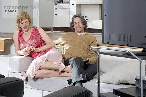 Eine Frau  die ein Handy neben einem Mann benutzt  der fernsieht.
