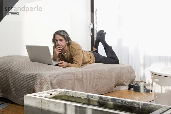Ein Mann  der auf einem Bett liegt und einen Laptop benutzt.