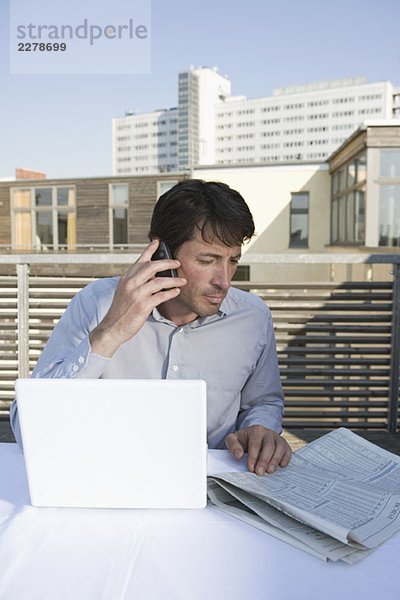 Ein Geschäftsmann sitzt auf einer Dachterrasse mit Zeitung  Handy und Laptop.