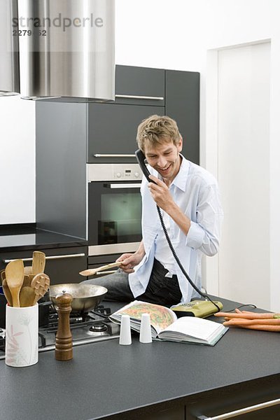 Ein Mann  der auf einer Küchenarbeitsplatte sitzt und das Telefon benutzt.