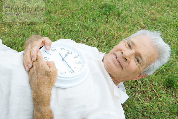 Älterer Mann auf Gras liegend  Uhr haltend  lächelnd vor der Kamera