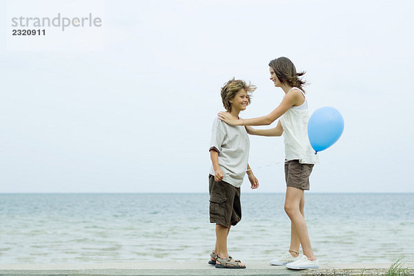 Geschwister stehen von Angesicht zu Angesicht am Strand  Mädchenhände auf den Schultern des Bruders  Junge hält Ballon