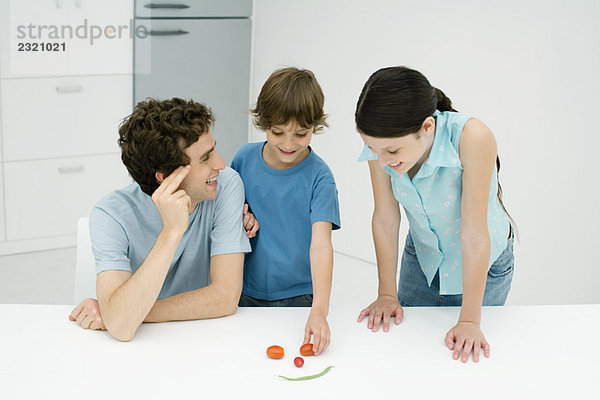 Vater und zwei Kinder in der Küche  die aus Gemüse ein Smiley machen.