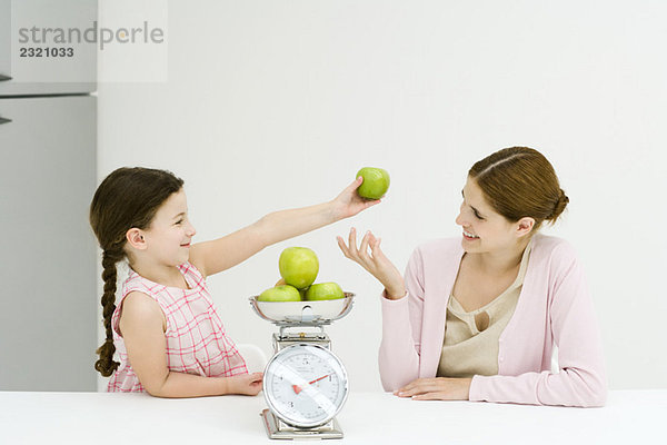 Mutter und Tochter wiegen Äpfel auf der Waage  lächeln sich an  Mädchen hält einen Apfel hoch.