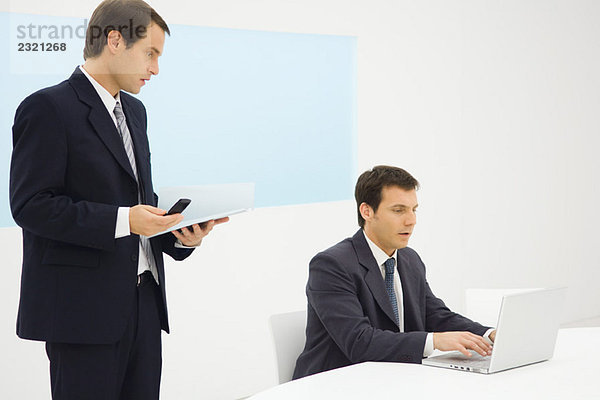 Zwei Geschäftsleute im Büro  die zusammen auf den Laptop schauen  einer hält das Handy.
