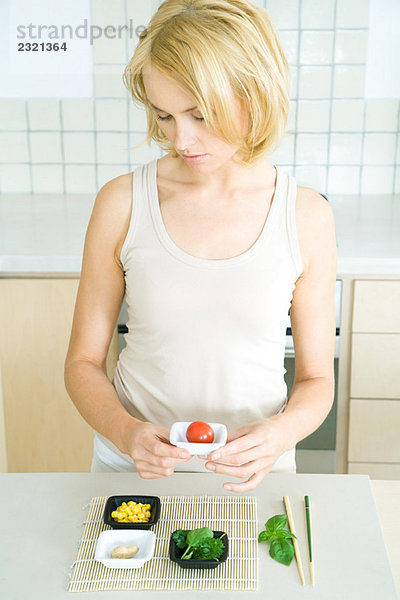 Junge Frau steht in der Küche  hält Kirschtomate in kleiner Schüssel  schaut nach unten.