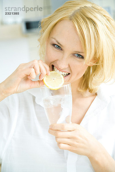 Frau hält ein Glas Wasser  beißt in die Zitronenscheibe  schaut in die Kamera