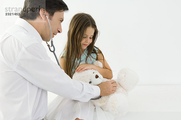 Arzt sitzt mit kleinem Mädchen  untersucht Teddybär mit Stethoskop