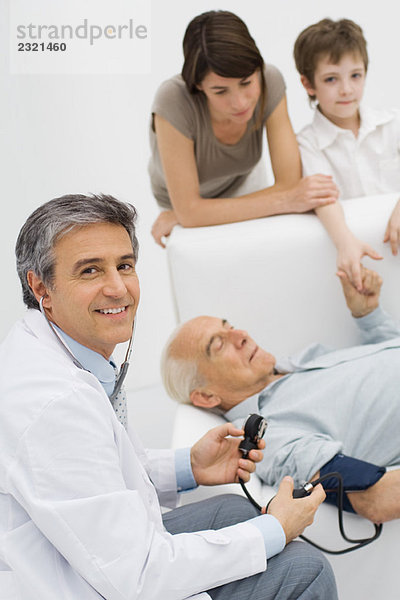 Arzt misst den Blutdruck des Patienten  lächelt über die Schulter vor der Kamera  Familie des Patienten im Hintergrund