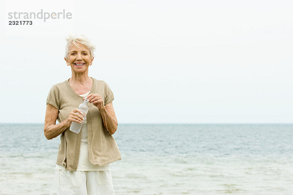 Seniorin steht am Strand  hält eine Flasche Wasser  lächelt die Kamera an.