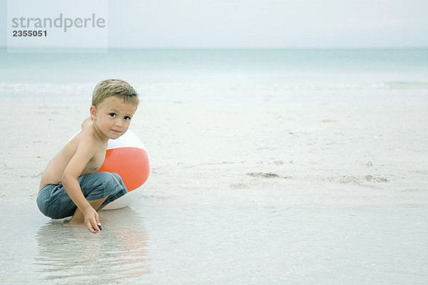 Kleiner Junge kauert im Wasser am Strand  spielt mit dem Strandball  schaut in die Kamera