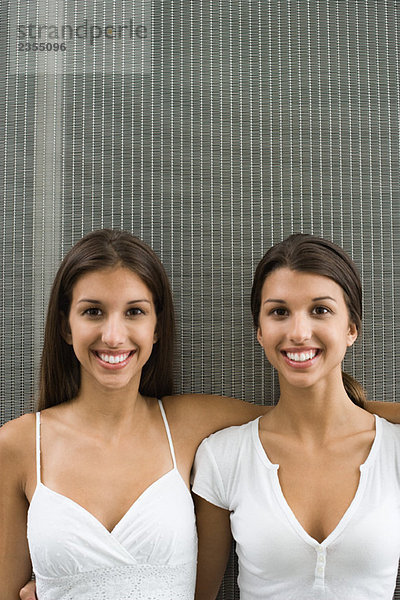 Teenager-Zwillingsschwestern mit Armen umeinander  beide lächelnd vor der Kamera  Porträt