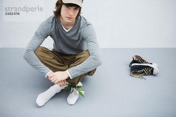 Junger Mann sitzt auf dem Boden neben seinen Schuhen  hält die Blume und schaut in die Kamera.