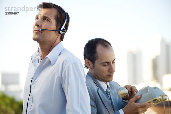 Zwei Männer stehen Rücken an Rücken  einer mit Headset  der andere mit Festnetztelefon.
