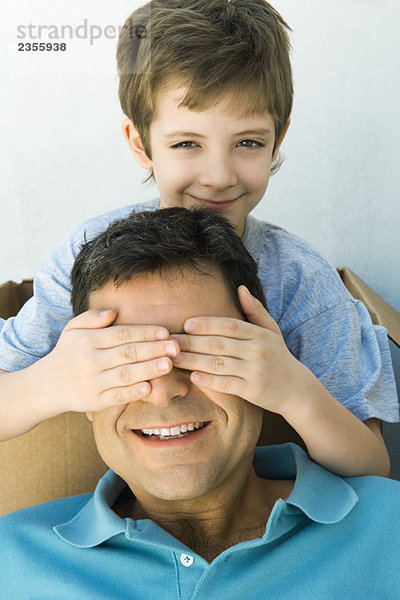Junge mit den Händen über den Augen seines Vaters  mit Blick auf die Kamera  beide lächelnd