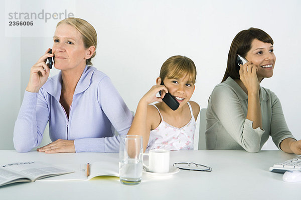 Mutter und zwei Töchter sitzen am Tisch  jede mit dem Handy  lächelnd.