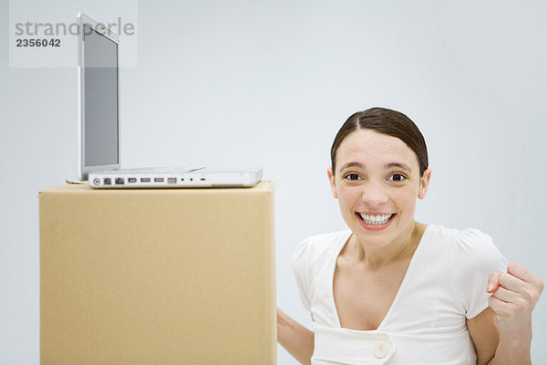 Junge Frau mit Laptop-Computer auf dem Karton  lächelnd in die Kamera  Fäuste zusammendrückend