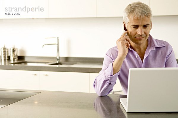 Älterer Mann  der am Laptop arbeitet  während er mit dem Handy spricht