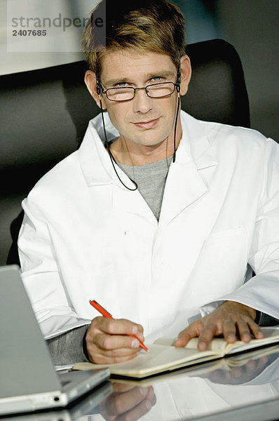 Porträt eines männlichen Arztes  der an einem Schreibtisch in seinem Büro sitzt und in ein Notizbuch schreibt.