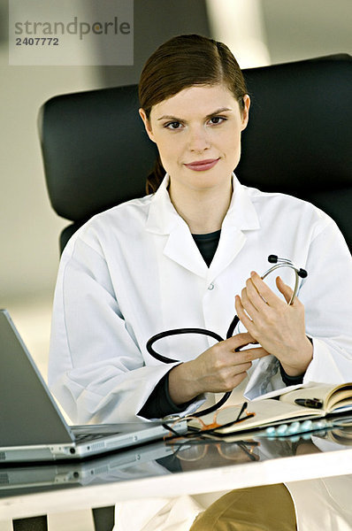Porträt einer Ärztin  die am Schreibtisch sitzt und ein Stethoskop im Büro hält.