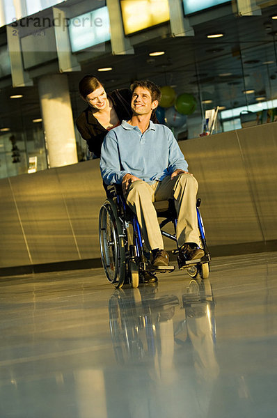 Junge Frau  die einen männlichen Patienten im Rollstuhl schiebt.