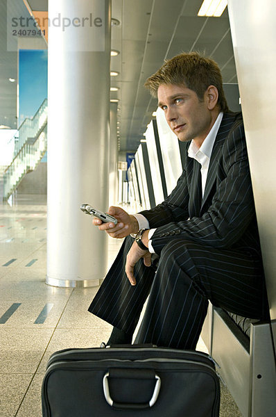 Geschäftsmann  der in einer Flughafenlounge sitzt und ein Handy bedient.